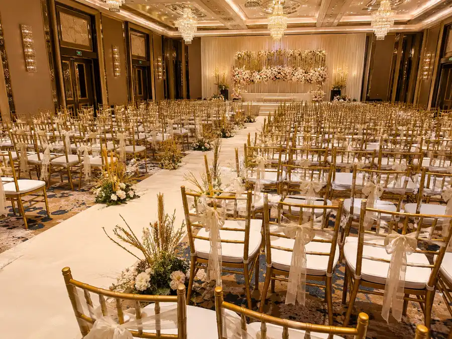 Gold Chiavari chairs arranged for a ballroom wedding ceremony with white aisle and floral stage